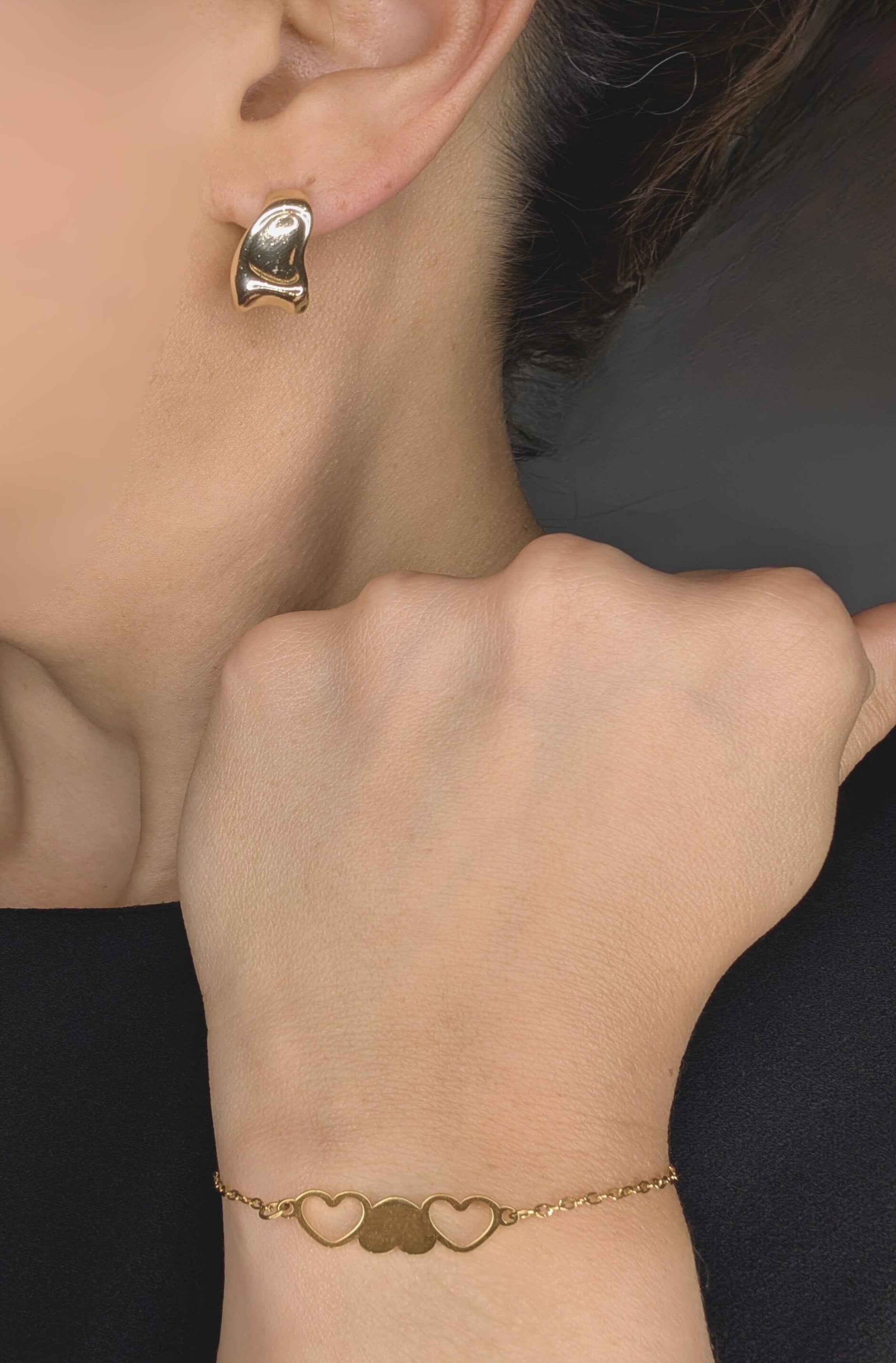 Gold heart-shaped earring and bracelet on a person against a dark background